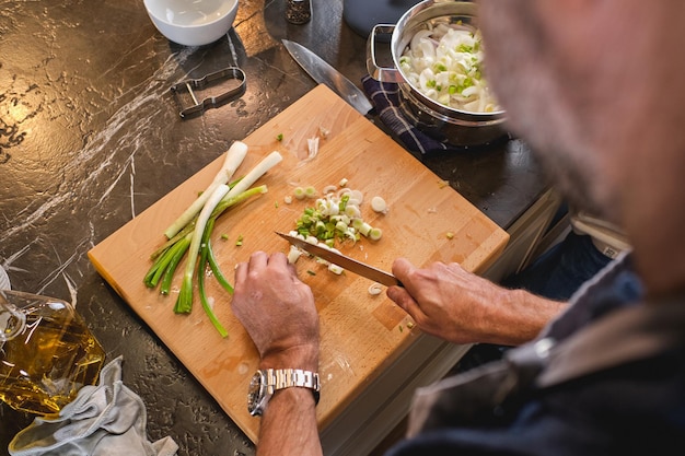 From above of crop anonymous male cook cutting fresh green onion with knife on chopping board at counter in light kitchen