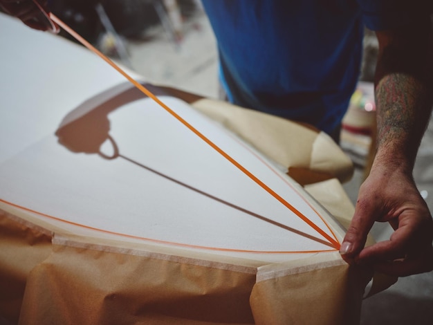 From above of crop anonymous male master putting decorative element on white handmade surfboard surface while making board design in workshop