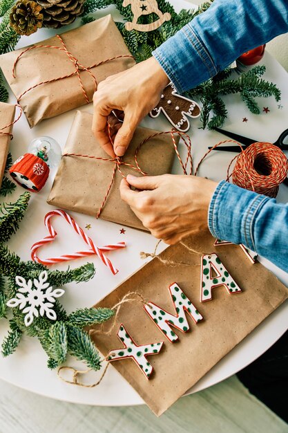 From above of crop unrecognizable person wrapping Christmas presents in paper with rope at table with fir branches and glass snow globe near letters and candy canes