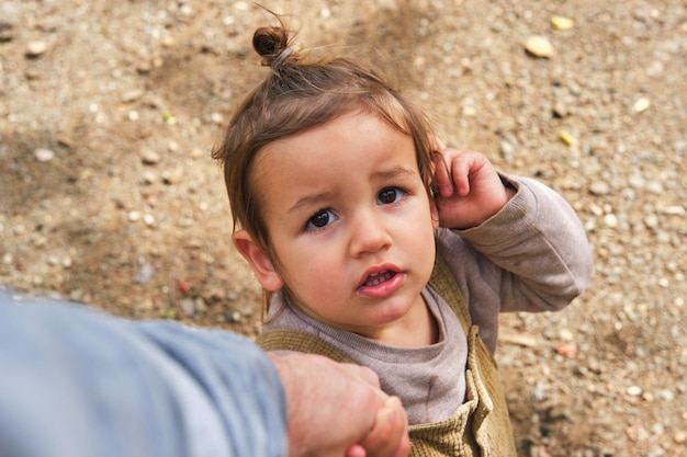 From above cute little girl holding parents hand and touching ear while standing on dirt road and looking at camera