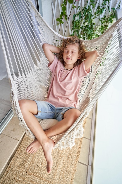 From above full length of adorable barefoot boy in casual outfit relaxing in wicker hammock with closed eyes and crossed legs on balcony with rug