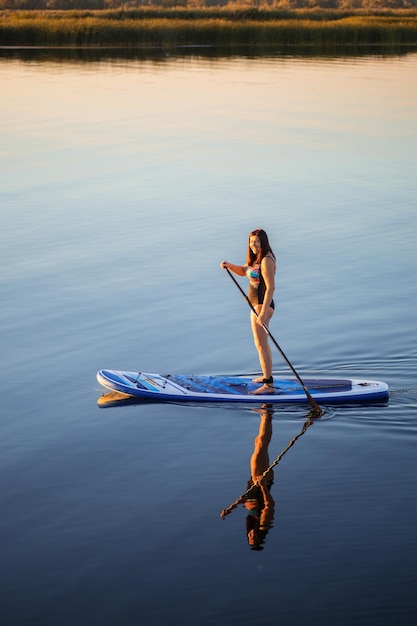 From above portrait of woman of middle age paddle boarding on lake with her clear reflection in water in evening time with reeds in background Active lifestyle