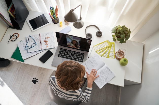 From above of schoolboy with exercise book doing homework during video chat with classmate on laptop at table with notebooks