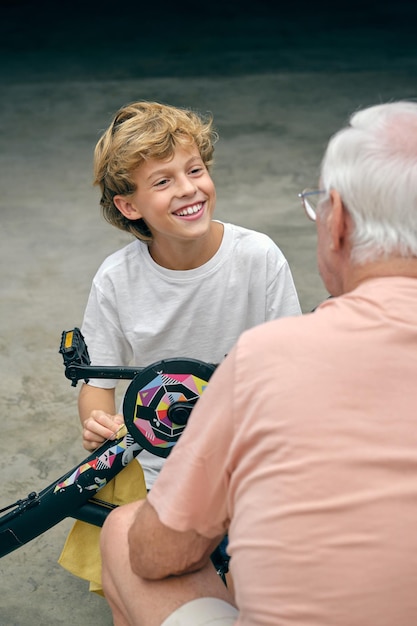 From above of smiling grandson sitting with bicycle and looking at crop grandfather with love