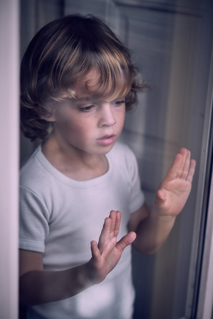 From above through glass of curious little boy with curly blond hair in white t shirt looking thought window at home