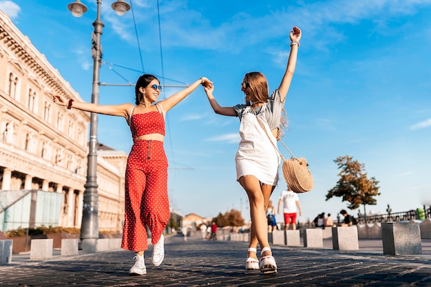 From below view of female friends holding hands and walking on old pavement along city street