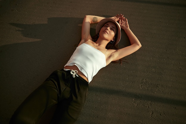 Photo from above of young hipster tattooed female in trendy summer outfit and hat, lying with eyes closed on asphalt street while enjoying sunny summer day in city