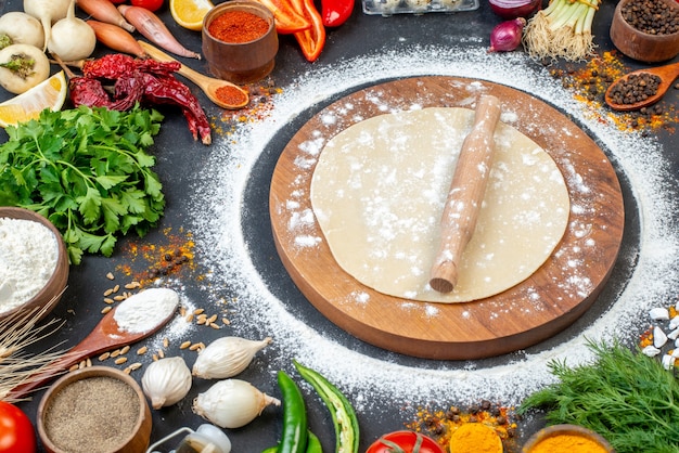 Front close view of circle dough rolling-pin on a wooden board with flour around and fresh vegetables spices on black surface