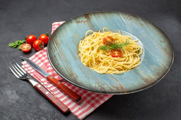 Front close view of delicious spagetti served with tomatoes greens and cutlery set on red stripped towel on black background