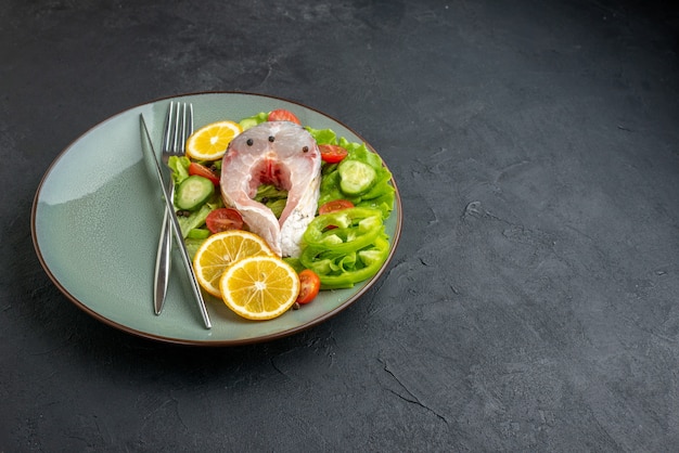 Front close view of raw fish and fresh vegetables lemon slices and cutlery set on a gray plate on the right side on black surface with free space