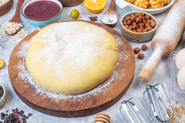 Front close view of raw pastry flour on round board grater fresh fruits jam cookies hazelnuts on stained white background