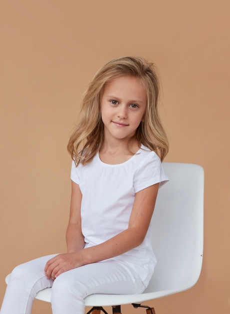 Front portrait of pretty smiling little girl with wavy blond hair, dressed in white clothes, sitting on whait chair