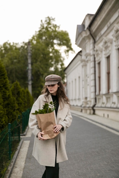 Front portrait of a stylish young woman with a paper pack of green leaf plant on urban city. Woman walking in cold day. Fashion, lifestyle concept.