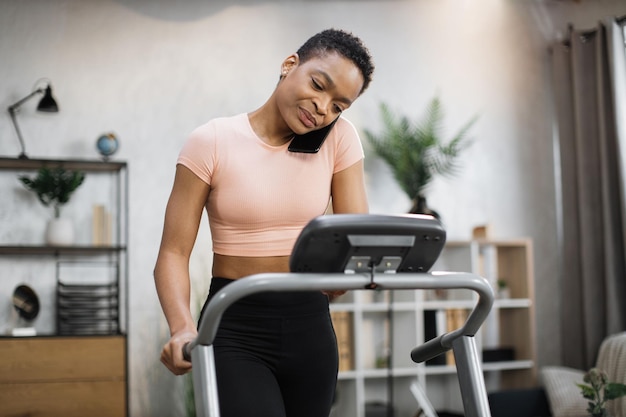 Front view of african sporty woman in pink tshirt working out in treadmill while having phone call