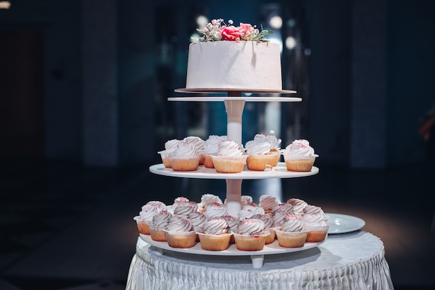 Front view of beautiful wedding cake with flowers staying on table. Delicious dessert for celebration. Lovely pair at background. Concept of love, confectionery and biscuit.