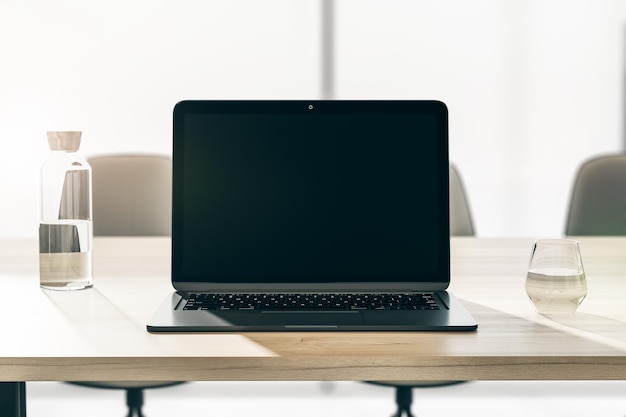 Front view on blank black modern laptop screen with space for your logo or text on wooden table with glass and bottle of water on sunlit office background 3D rendering mock up