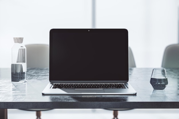 Front view on blank modern laptop screen with space for your logo or text on dark table with glass and bottle of water on sunlit office background 3D rendering mockup