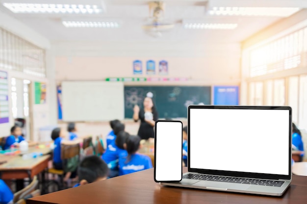 Front view blank screen of black digital laptop and smartphone with tablet on the wood table Children students in classroom background