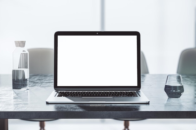 Front view on blank white modern laptop screen with space for your logo or text on dark concrete table with glass and bottle of water on sunlit office background 3D rendering mock up