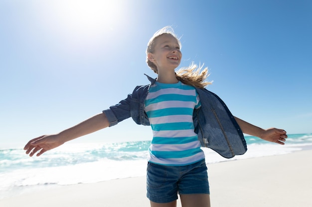 Front view of a Caucasian girl on a sunny beach, smiling with arms outstretched, with blue sky and sea in the background