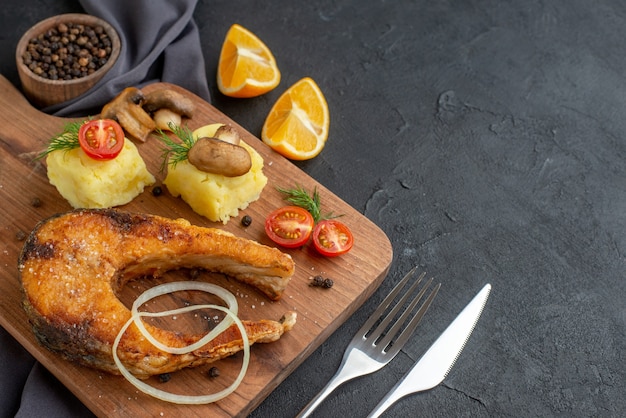 Front view of delicious fried fish meal with mushrooms vegetables cheese on wooden board lemon slices pepper on dark color towel cutlery set on the right side on black distressed surface