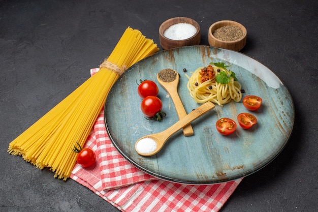 Front view of delicious spagetti served with meat greens tomatoes salt pepper on a plate and raw pasta on red stripped towel on black background