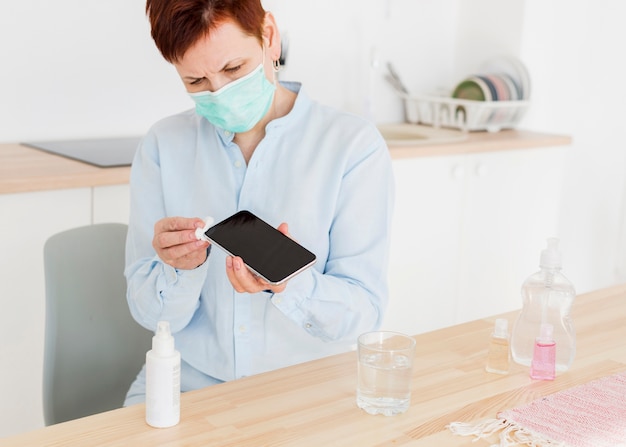 Front view of elder woman with medical mask disinfecting her smartphone at home