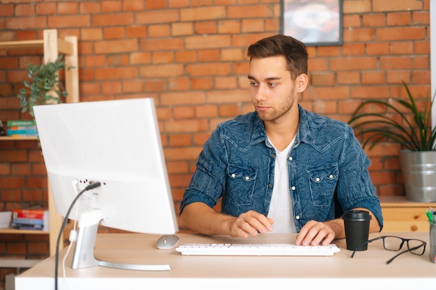 Front view of focused handsome young freelance programmer male working on desktop computer sitting at desk at home office