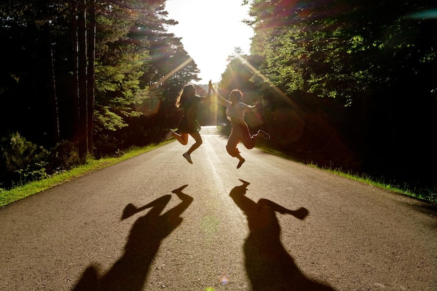 Front view of friends jumping in the middle of the road. Horizontal view of women jumping in backlighting shadows traveling in nature background. Summer holidays concept.