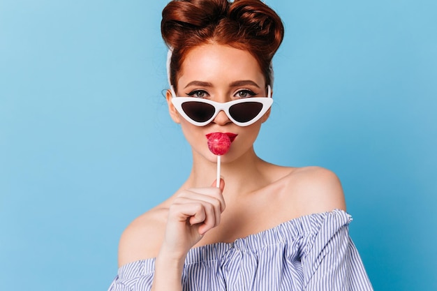 Front view of ginger girl in sunglasses licking candy Confident pinup woman holding lollipop on blue background