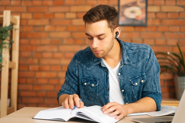 Front view of handsome focused young man reading business paper book sitting at desk with laptop at home office