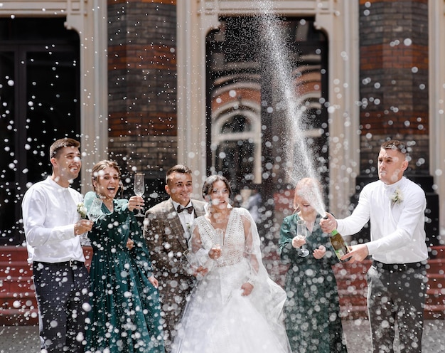 Front view joyful newlyweds with friends standing together and holding crystal wineglasses laughing and looking to boy which opening bottle and making champagne splash during wedding walk in open air