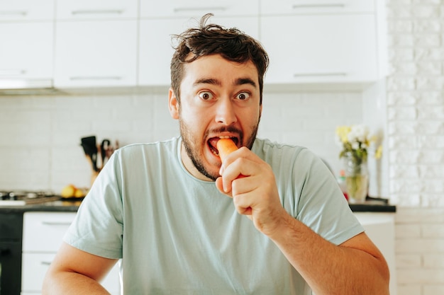 Photo front view looking at camera a young man sits at a table and eats a raw carrot with his eyes wide