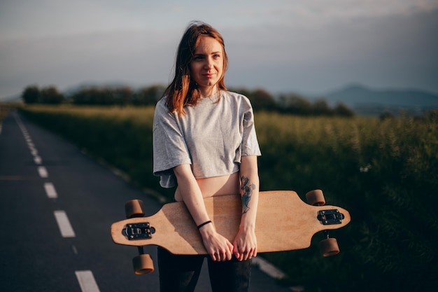 Front view low angle Funny young girl in jeans and top stands with big longboard Green nature background