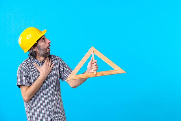 Front view male builder in helmet holding triangle wooden figure on blue background constructor design engineer architecture building job color