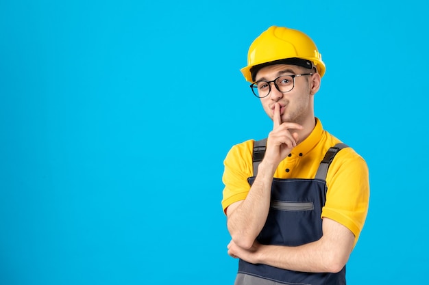 Front view male builder in uniform and helmet asking to be quiet on blue 