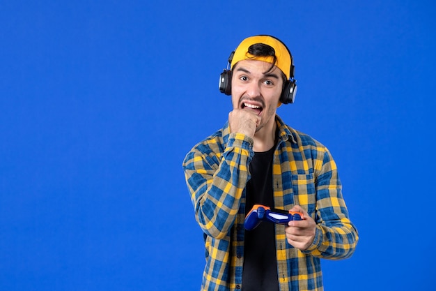 Front view of male gamer with gamepad and headphones playing video game on blue wall