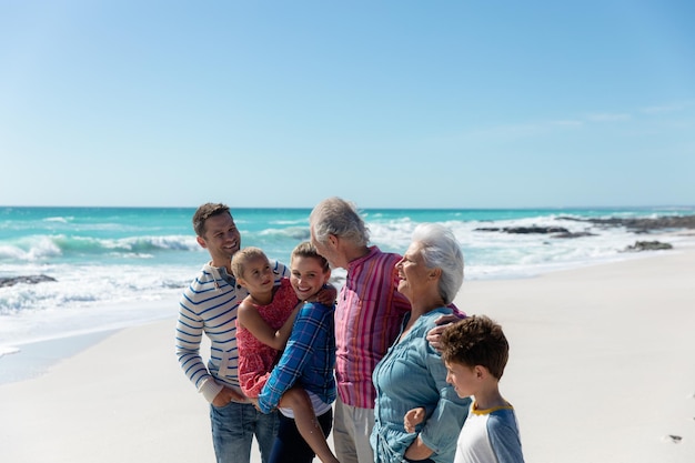 Front view of a multi-generation Caucasian family standing on the beach with blue sky and sea in the background, embracing and smiling