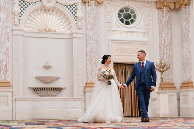 Front view o cute and pretty girl in long wedding dress holding flowers arrangement smiling and looking at bridegroom while walking with him inside of luxury hall