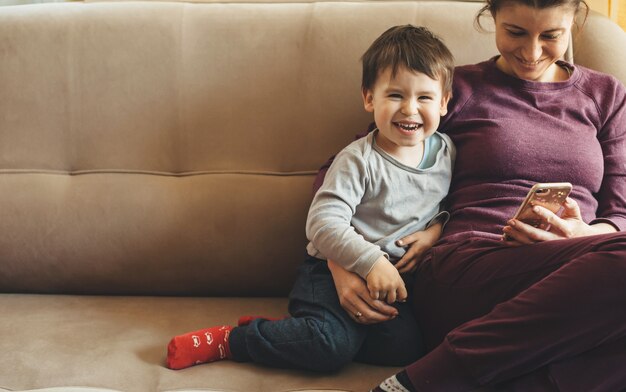 Front view portrait of a caucasian mother and son sitting on the sofa and using a mobile while smiling