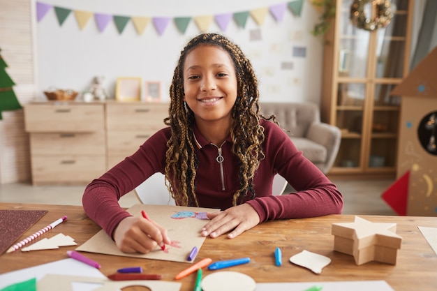 Front view portrait of cheerful African-American girl looking at camera while enjoying drawing sitting at desk in home interior, copy space