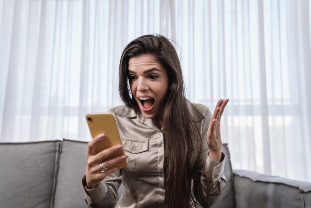 Front view portrait of an excited Latin woman holding phone looking at you sitting on a sofa