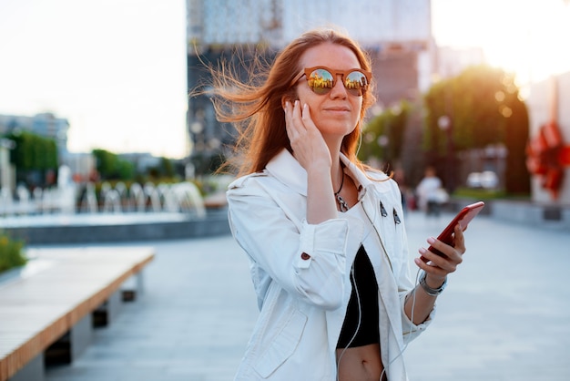 Front view portrait modern fashion happy woman hipster walking and using a smart phone on a city street wearing sunglasses in the summer sun. Internet, online service, Telephone, girl, female