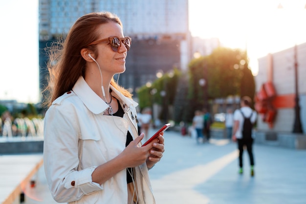 Front view portrait modern fashion happy woman hipster walking and using a smart phone on a city street wearing sunglasses in the summer sun