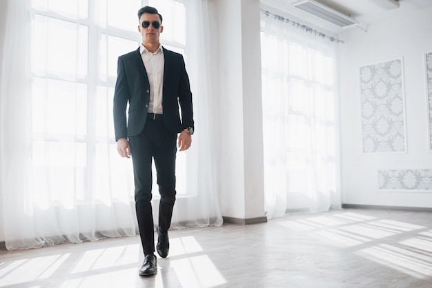 Front view. Portrait of young stylish man in classic business wear walking in the white room.