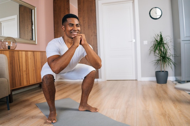 Front view of smiling AfricanAmerican man exercising and doing squats online in front of laptop