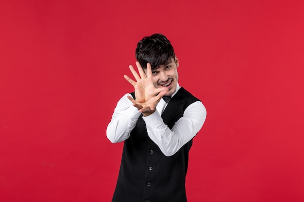 Front view of smiling male waiter in a uniform with butterfly on neck making exact something on red wall