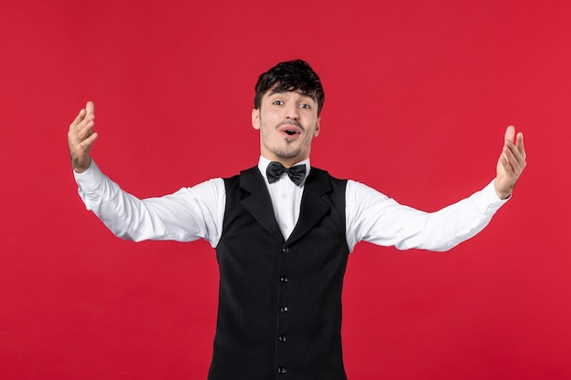 Front view of smiling wondering male waiter in a uniform with butterfly on neck and making exact something on red wall