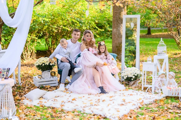 Front view of stylish parents and children smiling and looking at camera while sitting on comfortable sofa in park which decorated by beautiful decor and potted flowers during autumn day