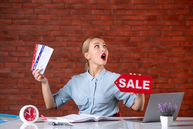 front view of travel agent sitting behind her working place holding tickets and sale nameplate manager indoors agency occupation assistant tourism map